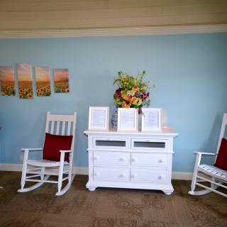 A cozy interior setting featuring a white dresser adorned with decorative flowers, flanked by two rocking chairs against a light blue wall with artwork.