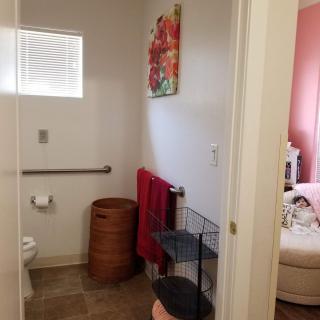 A view of a bathroom featuring a toilet, towel rack, and laundry basket, with a painting on the wall and a glimpse into an adjoining room.