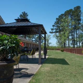 A backyard patio with a gazebo and outdoor furniture, overlooking a well-maintained lawn and trees under a clear blue sky.