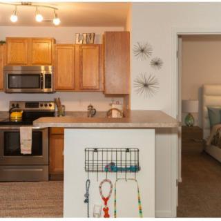 A modern apartment kitchen with wooden cabinets and stainless steel appliances, opening into a cozy living area.