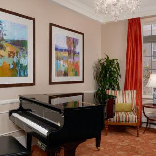 A stylish living room features a grand piano, colorful artwork on the walls, an elegant chandelier, and a comfortable armchair near the window with red curtains.