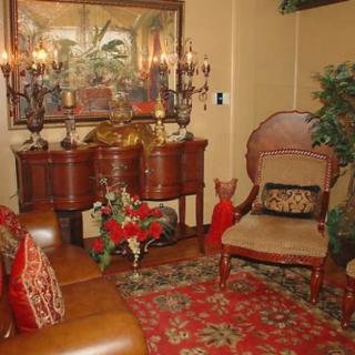 A warmly decorated room featuring traditional furniture and vintage decor, with an armchair, a sideboard adorned with candles, and a vibrant floral arrangement on a patterned rug.