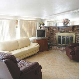 A cozy living room featuring a comfortable sofa, armchair, TV, and a brick fireplace with large windows and blinds.