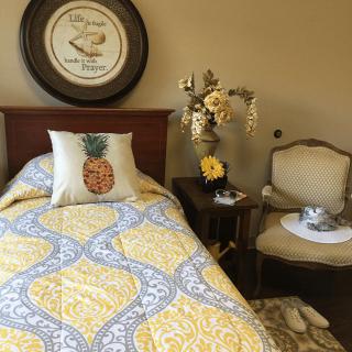 A cozy bedroom with a yellow and gray bedspread, pineapple-themed decor, and a floral arrangement on a side table.