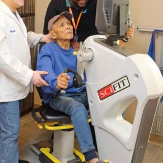 An elderly man receiving assistance while using an exercise machine for physical therapy.