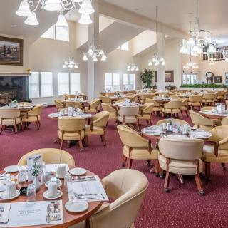 A spacious restaurant dining room with elegant chandeliers and neatly arranged tables and chairs on red carpet flooring.