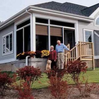 An elderly couple with their dog standing on the porch of their house, surrounded by a well-maintained garden.