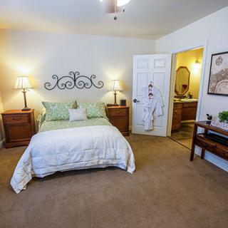 A cozy bedroom featuring a bed with green and white linens, flanked by wooden nightstands and lamps. The room includes carpet flooring, decorative wall art, and a view into an adjoining bathroom.