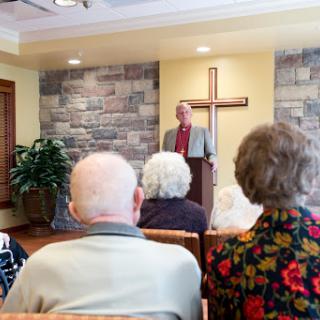 A speaker addresses a group of elderly attendees in a church setting, with a cross prominently displayed in the background.
