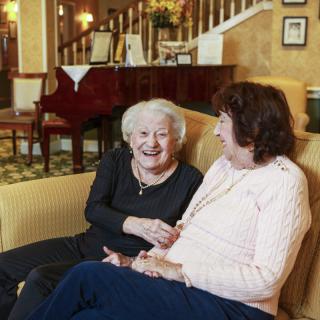 Two elderly women sharing a laugh while sitting on a sofa indoors.