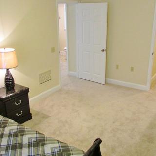 A cozy bedroom with a nightstand and lamp, carpeted flooring, and an open doorway.
