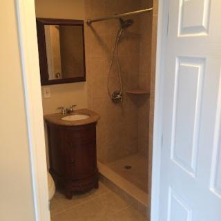 A view of a small bathroom with a shower, sink, and mirror through an open doorway.