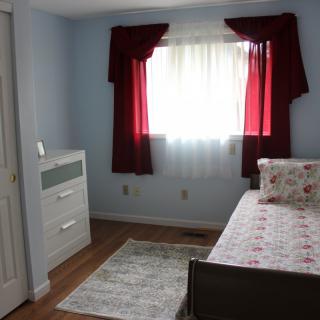 A cozy bedroom featuring a single bed with floral bedding, a white dresser, and red curtains over a window.