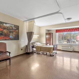 A clean and spacious hospital room featuring a bed, chair, window with blinds, and artwork on the wall.