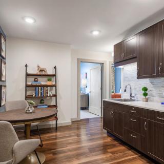 A modern kitchen with wooden flooring and dark cabinets, featuring a cozy dining area and a bookshelf.