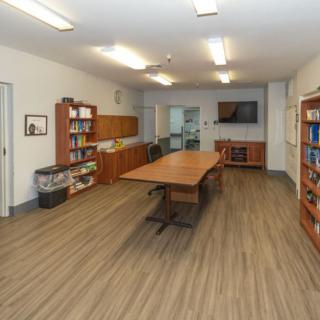 A modern meeting room with a large conference table, bookshelves, and a wall-mounted television.