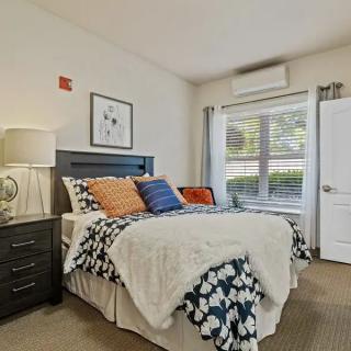 A cozy bedroom with a bed adorned with pillows and a bedside table, illuminated by natural light from a large window.