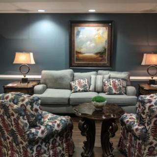 A cozy living room setting featuring a sofa with decorative cushions, two patterned armchairs, and a central coffee table. Two lamps provide lighting on either side of the sofa, with a landscape painting on the wall.