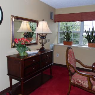 A cozy living room corner featuring a wooden dresser with a lamp and mirror, potted plants on the windowsill, and an elegant chair.