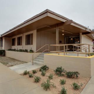 A single-story building with a ramp and stairs leading to the entrance, surrounded by landscape plants.