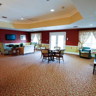 A spacious living room with comfortable furnishings, including a sofa, chairs, and a table. The room is well-lit with natural light coming through the windows and features a television mounted on the wall.