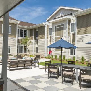 A sunny outdoor patio area with tables, chairs, and umbrellas in front of a residential building.