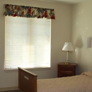 A simple bedroom with a bed, window with blinds and curtains, nightstand, and lamp.