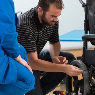 A professional performs maintenance on a wheelchair while discussing with a colleague.