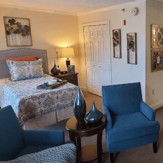 A cozy bedroom with a patterned bedspread, two blue chairs, and tasteful artwork on the walls.