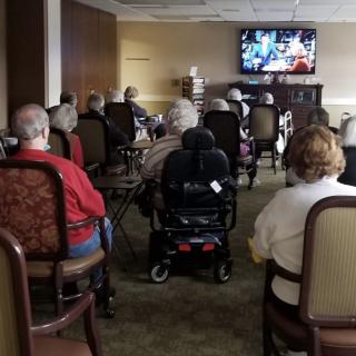 A group of elderly people watching television in a community room, with one person in a wheelchair.