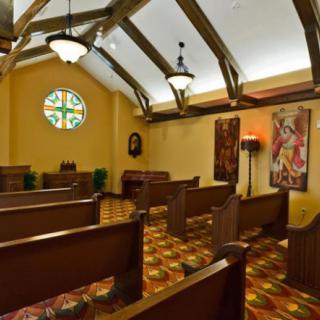 A serene chapel interior with wooden pews, stained glass window, and religious artwork.