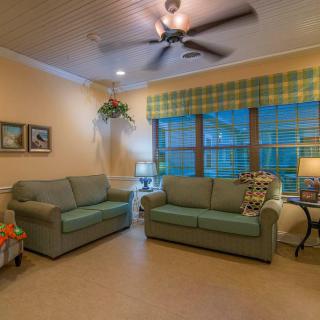 A cozy living room with two sofas, a ceiling fan, large windows with plaid curtains, and warm lighting from lamps.