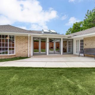 A single-story house with a spacious backyard and patio area, featuring large windows and a well-maintained lawn under a partly cloudy sky.