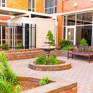 A peaceful courtyard featuring a fountain, surrounded by greenery and benches, adjacent to a brick building.