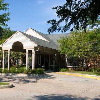 An exterior view of a building entrance with columns and a surrounding driveway, shaded by trees.