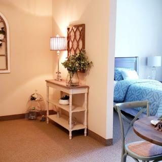 A cozy interior featuring a bedroom with blue bedding, a side table with a lamp and plants, and warm-toned walls.