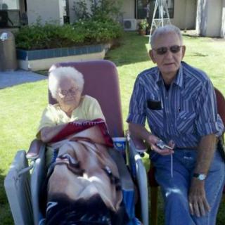 An elderly couple enjoying a sunny day outdoors, with one seated in a wheelchair and covered with a blanket.