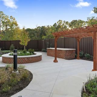 A beautifully landscaped outdoor area featuring a pergola, paved walkway, and a garden with trees and plants. A tall wooden fence provides privacy.