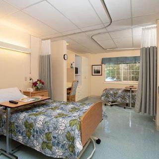 A well-furnished hospital room with two beds, floral bedspreads, a window, and privacy curtains.