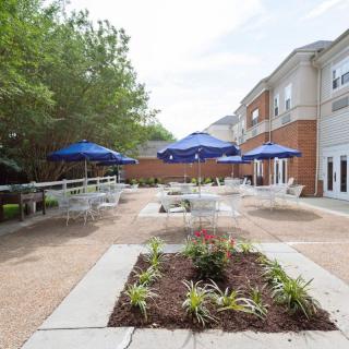 An outdoor patio area with tables and blue umbrellas, surrounded by greenery and a building.