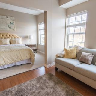 A cozy and inviting bedroom featuring a queen-sized bed with a soft yellow comforter and a comfortable sofa, illuminated by natural light from large windows.