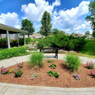 A scenic garden with a bench surrounded by lush greenery and colorful plants under a blue sky.