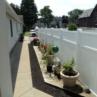 A neat row of potted plants lines a pathway next to a white fence in a residential area.