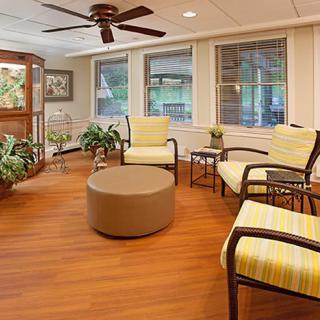 A cozy indoor seating area featuring comfortable chairs, a circular ottoman, and lush indoor plants against large windows.