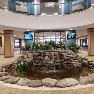 A modern indoor lobby featuring a central stone fountain surrounded by plants.