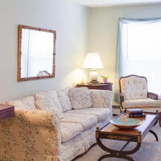 A cozy living room with a patterned sofa, armchair, and a coffee table, illuminated by a lamp next to a large window.