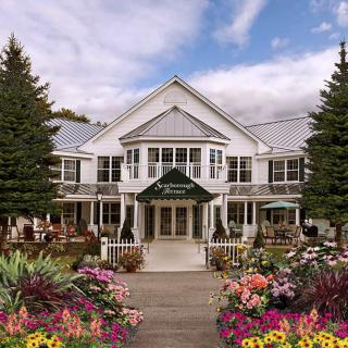 A charming white building with a welcoming entrance, surrounded by vibrant flowers and lush trees under a partly cloudy sky.