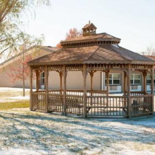 A wooden gazebo in a sunny outdoor setting, surrounded by autumn foliage.