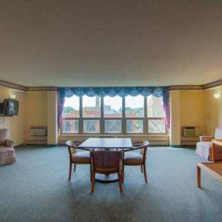 A spacious living room with large windows, carpeted floor, and a central table surrounded by chairs.