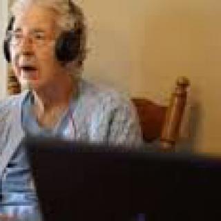 An elderly woman wearing headphones, sitting indoors and engaging with a computer.